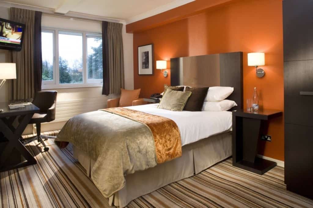 A classy bedroom that uses colorful striped floor carpet, dark furniture and a large cabinet. The head side wall is red orange while the other walls and roof are white.